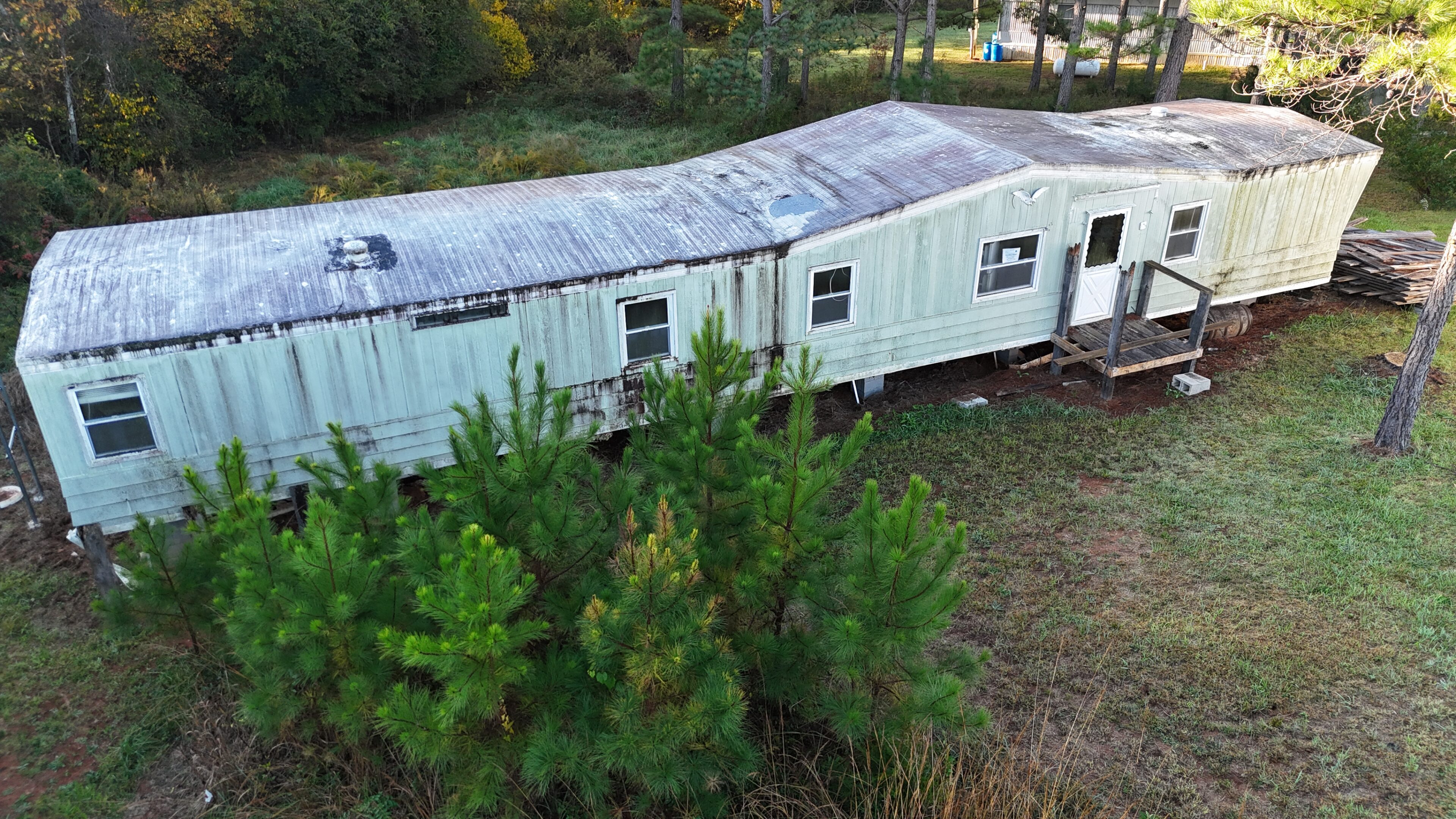 White mobile home with a rusty roof surrounded by greenery.