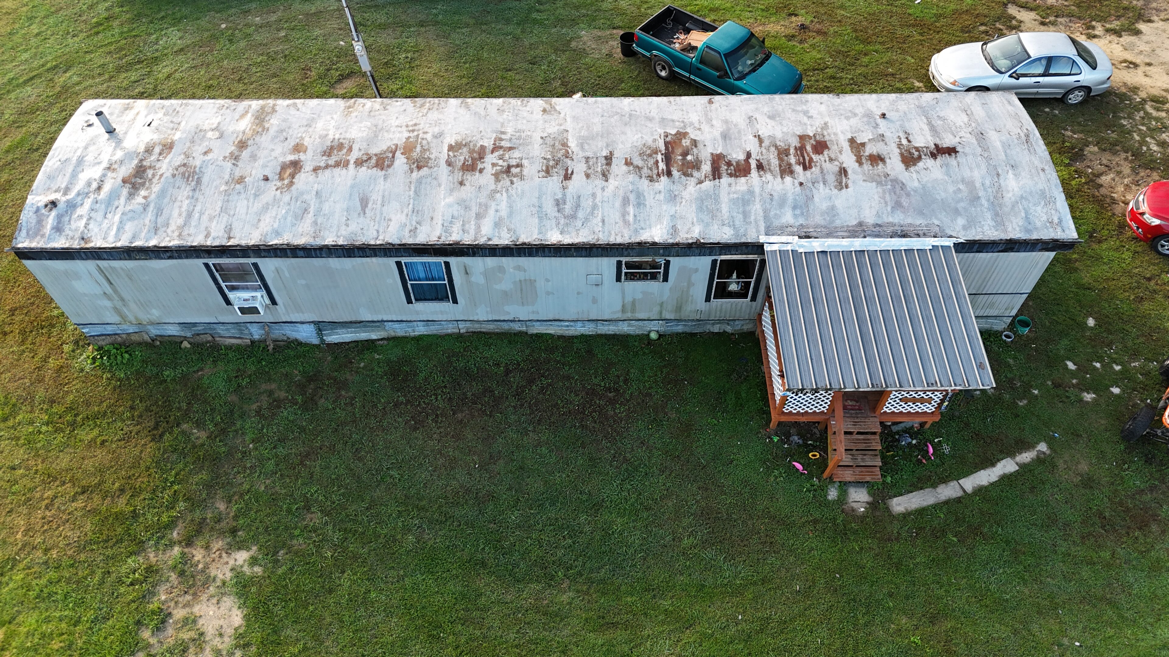 Aerial view of a weathered mobile home with a rusty roof and a blue car nearby.