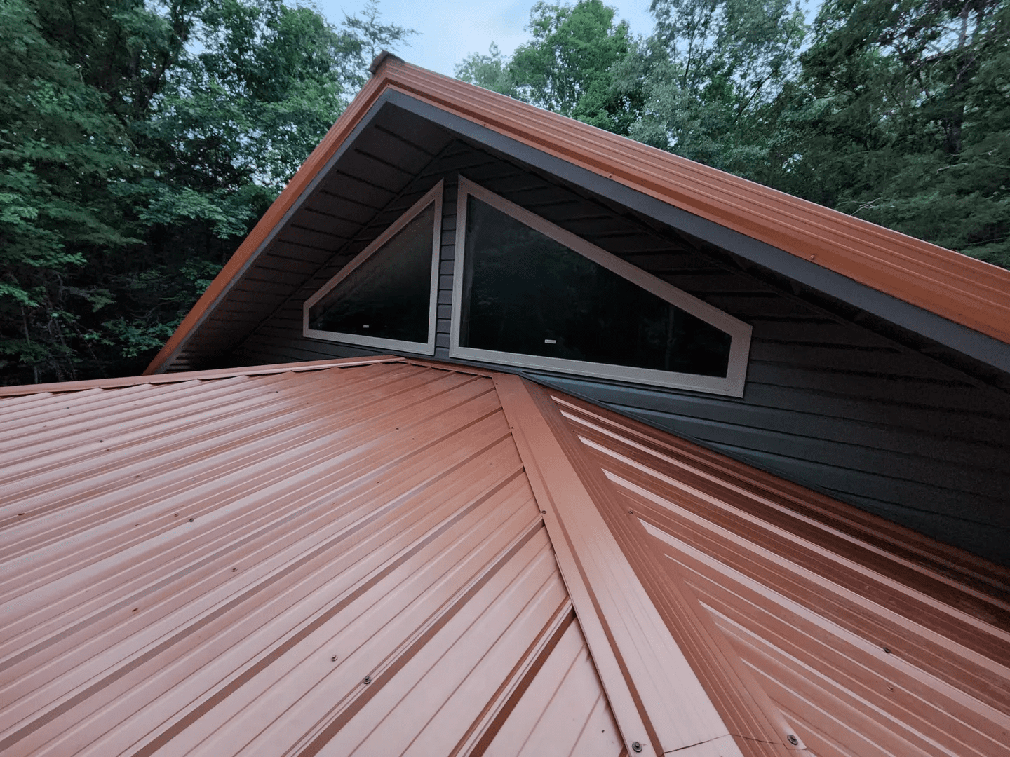 View of a brown metal roof with triangular windows and surrounding trees.